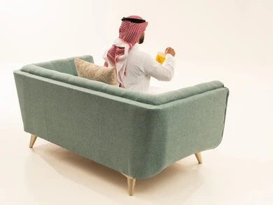 Enjoying popcorn, home entertainment activities, a portrait from behind of a Saudi Arabian Gulf man wearing traditional attire and a ghutrah sitting on the sofa holding a box of corn, delicious snacks and crisps, eating light and healthy snacks, enjoying eating popcorn, entertaining oneself and having a good time, white background.