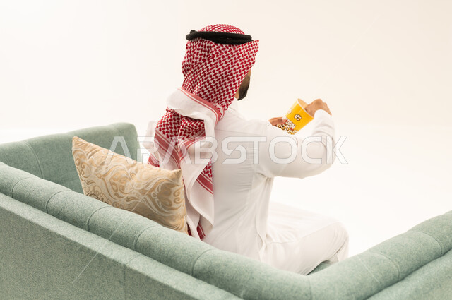 انشطة ترفيهية منزلية، الاستمتاع بتناول الفشار، بورتريه من الخلف لرجل عربي خليجي سعودي يرتدي الثوب التقليدي و الشماغ يجلس على الاريكة يحمل بيده علبة الذرة، سناكات ومقرمشات لذيذة، تناول سناكات خفيفة وصحية، الاستمتاع باكل البوب كورن، الترفيه عن النفس وقضاء وقت ممتع،خلفيه بيضاء