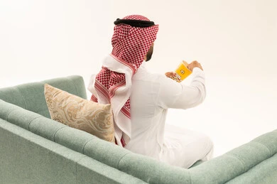 انشطة ترفيهية منزلية، الاستمتاع بتناول الفشار، بورتريه من الخلف لرجل عربي خليجي سعودي يرتدي الثوب التقليدي و الشماغ يجلس على الاريكة يحمل بيده علبة الذرة، سناكات ومقرمشات لذيذة، تناول سناكات خفيفة وصحية، الاستمتاع باكل البوب كورن، الترفيه عن النفس وقضاء وقت ممتع،خلفيه بيضاء