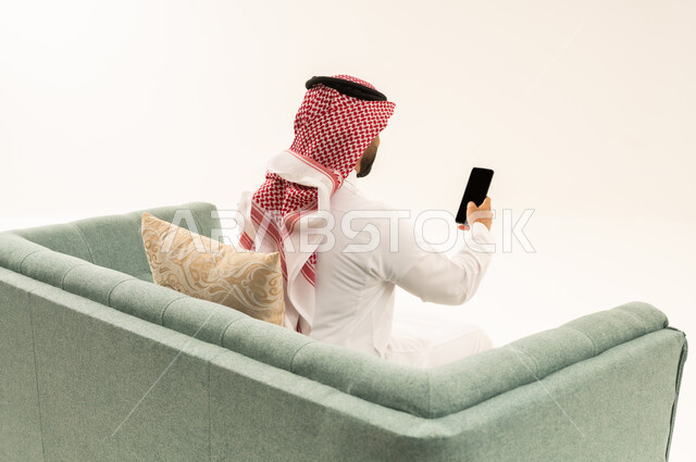 تصفح مواقع التواصل الاجتماعي عبر الانترنت، تطور التكنولوجيا والتقنيات الحديثة، بورتريه من الخلف لرجل عربي خليجي سعودي يرتدث الثوب التقليدي و الشماغ يجلس على الاريكة يقوم باستخدام الهاتف المحمول، إجراء مكالمات فيديو عبر الجوال، خلفية بيضاء