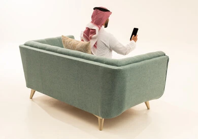 Following the latest news on social media, browsing social media sites, electronic marketing via smart devices, a portrait from behind of an Arab Saudi Gulf man wearing a ghutrah and traditional thobe sitting on a sofa using a mobile phone, white background.