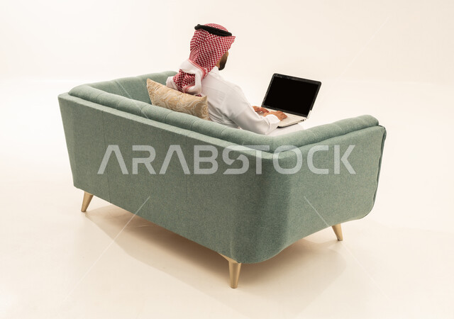 تصفح الانترنت ووسائل التواصل الاجتماعي، انجاز الاعمال والمهام عن بعد عبر الحاسب المحمول، بورتريه من الخلف لرجل عربي سعودي خليجي يرتدي الشماغ و الزي التقليدي يجلس على الاريكة يقوم بالعمل على جهاز الاب توب، خلفية بيضاء