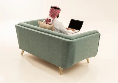 Browsing the internet and social media, completing work and tasks remotely via a laptop, a portrait from behind of an Arab Saudi Gulf man wearing a ghutrah and traditional attire sitting on a sofa working on a laptop, white background.
