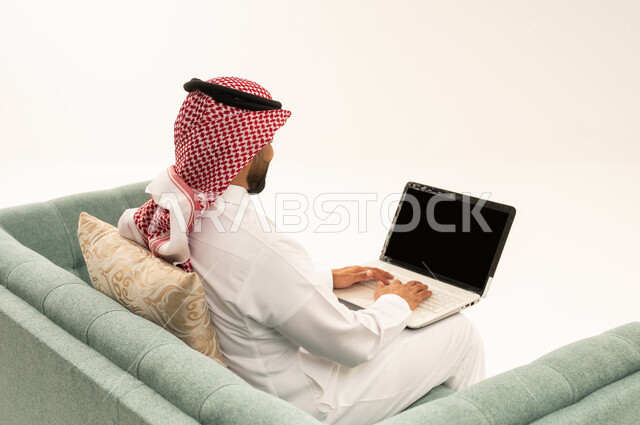انجاز الاعمال والمهام عن بعد عبر الحاسب المحمول، تصفح الانترنت ووسائل التواصل الاجتماعي، بورتريه من الخلف لرجل عربي سعودي خليجي يرتدي الشماغ و الزي التقليدي يجلس على الاريكة يقوم بالعمل على جهاز الاب توب، خلفية بيضاء