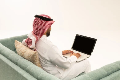 Completing tasks and assignments remotely via a laptop, browsing the internet and social media, a portrait from behind of an Arab Saudi Gulf man wearing a ghutrah and traditional attire sitting on a sofa working on a laptop, with a white background.