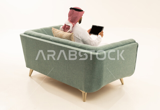 استعمال أجهزة تقنية حديثة، إنجاز المهام عن بعد، قضاء الوقت بتصفح برامج التواصل الاجتماعي، التسوق عبر الانترنت باستخدام التابلت، بورتريه من الخلف لرجل عربي سعودي خليجي يرتدي الشماغ و الزي التقليدي يجلس على الاريكة يستخدم الجهاز اللوحي، خلفية بيضاء