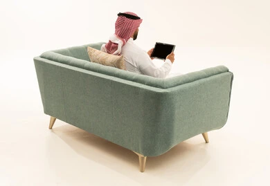 استعمال أجهزة تقنية حديثة، إنجاز المهام عن بعد، قضاء الوقت بتصفح برامج التواصل الاجتماعي، التسوق عبر الانترنت باستخدام التابلت، بورتريه من الخلف لرجل عربي سعودي خليجي يرتدي الشماغ و الزي التقليدي يجلس على الاريكة يستخدم الجهاز اللوحي، خلفية بيضاء