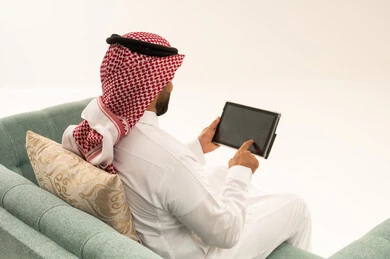 Completing tasks remotely, spending time browsing social media, shopping online using a tablet, using modern technological devices, a portrait from behind of an Arab Saudi Gulf man wearing a ghutrah and traditional attire sitting on a sofa using a tablet, white background.