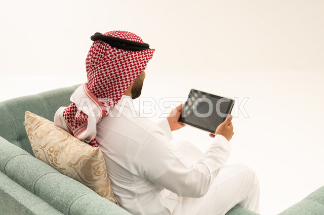 Spending time browsing social media, shopping online using a tablet, completing tasks remotely, using modern technological devices, a portrait from behind of a Saudi Arabian Gulf man wearing a ghutrah and traditional attire sitting on a sofa using a tablet, white background.