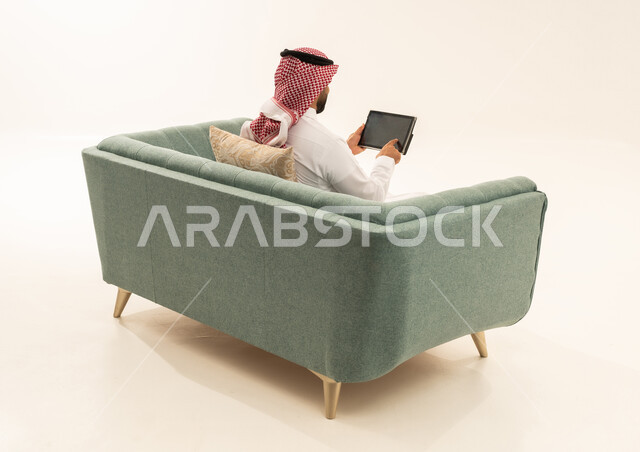 التسوق عبر الانترنت باستخدام التابلت, قضاء الوقت بتصفح برامج التواصل الاجتماعي, إنجاز المهام عن بعد، استعمال أجهزة تقنية حديثة، بورتريه من الخلف لرجل عربي سعودي خليجي يرتدي الشماغ و الزي التقليدي يجلس على الاريكة يستخدم الجهاز اللوحي، خلفية بيضاء