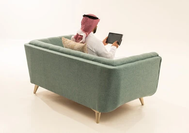 التسوق عبر الانترنت باستخدام التابلت, قضاء الوقت بتصفح برامج التواصل الاجتماعي, إنجاز المهام عن بعد، استعمال أجهزة تقنية حديثة، بورتريه من الخلف لرجل عربي سعودي خليجي يرتدي الشماغ و الزي التقليدي يجلس على الاريكة يستخدم الجهاز اللوحي، خلفية بيضاء