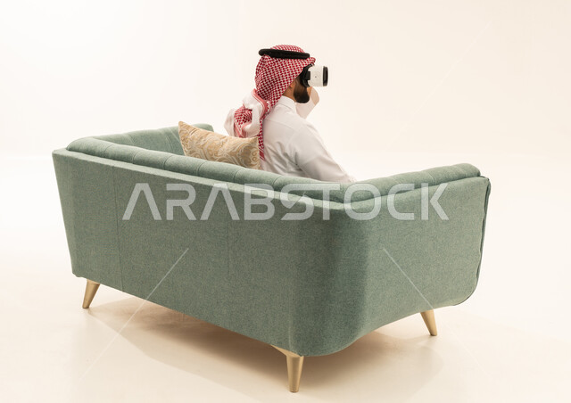 Integrating modern technologies and technology with fun and entertainment, simulating the imaginary world, a portrait from behind of a Saudi Arabian Gulf man wearing a white thobe and a ghutrah sitting on a sofa wearing 3D virtual reality glasses, gestures of immersion and focus, white background.