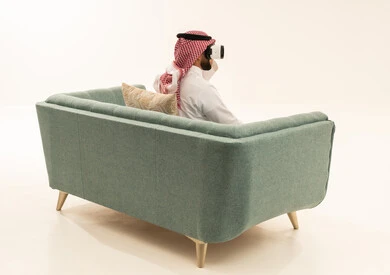 Integrating modern technologies and technology with fun and entertainment, simulating the imaginary world, a portrait from behind of a Saudi Arabian Gulf man wearing a white thobe and a ghutrah sitting on a sofa wearing 3D virtual reality glasses, gestures of immersion and focus, white background.