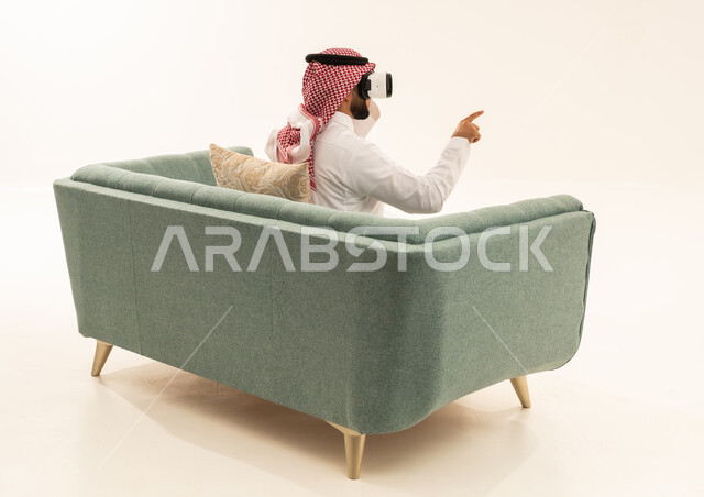 Referring to a place with expressions of integration and simulation of the imaginary world, attempting to click on something, a portrait from behind of a Saudi Arabian Gulf man wearing a white thobe and a ghutrah sitting on a sofa wearing 3D glasses raising his index finger upwards, white background.