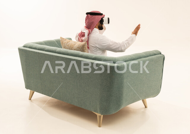 المتعة والتسلية بالمشاهدة عبر نظارة الواقع الافتراضي, ورتريه من الخلف لرجل عربي خليجي سعودي يرتدي الثوب الابيض و الشماغ يجلس على الاريكة يضع نظارات ثلاثية الأبعاد يقوم برفع وفتح كف يده للاعلى بتعابير الاندماج والتوقف عن فعل امر ما, خلفية بيضاء