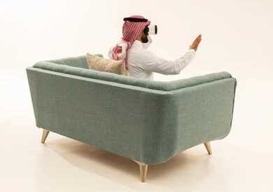 The enjoyment and entertainment of watching through virtual reality glasses, and a portrait from behind of a Saudi Arabian Gulf man wearing a white thobe and a ghutrah sitting on a sofa wearing 3D glasses, raising and opening his palm upwards with expressions of immersion and stopping from doing something, white background.