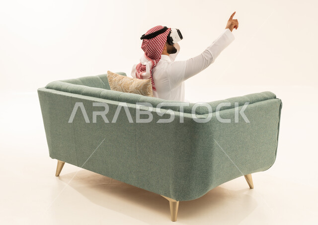 Attempting to click on something, a portrait from behind of a Saudi Arabian Gulf man wearing a white thobe and a ghutrah sitting on a sofa wearing 3D glasses, raising his index finger upwards, pointing to a place with expressions of immersion and simulating the imaginary world, white background.