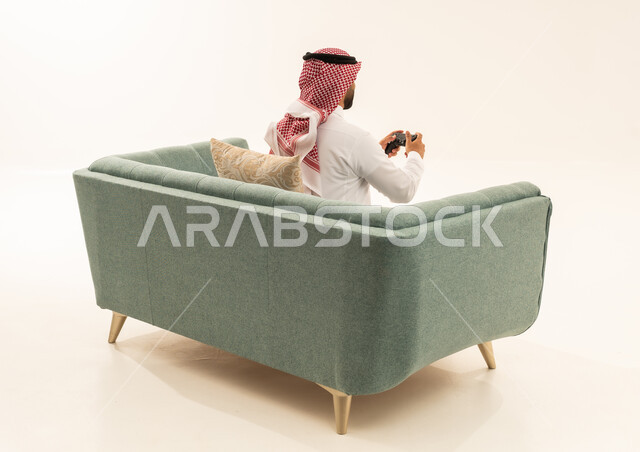 التفاعل والاندماج مع ألعاب البلايستيشن، تقنيات الكترونية ترفيهيه, بورتريه من الخلف لرجل عربي خليجي سعودي يرتدي الثوب الابيض و الشماغ يجلس على الاريكة يحمل بيده عصا التحكم، تجربة ترفيهية ممتعة ومثيرة في الأجهزة الحديثة، خلفية بيضاء