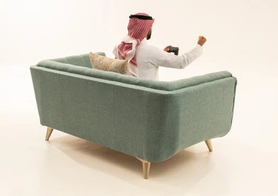Gestures of victory and winning, interaction and immersion with PlayStation games, entertaining electronic technologies, a portrait from behind of an Arab Gulf Saudi man wearing a white thobe and a ghutrah sitting on the sofa holding a game controller, an enjoyable and exciting entertainment experience on modern devices, white background.