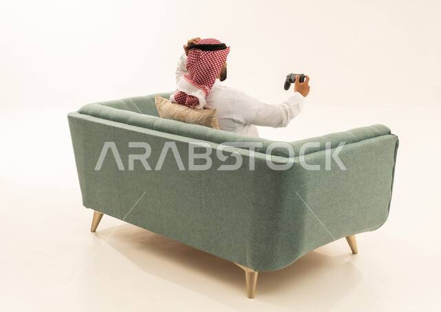 Entertainment and leisure with video games in free time, gestures of loss and disappointment, the use of electronic games and modern devices, a portrait from behind of a Saudi Arabian Gulf man wearing a white thobe and a ghutrah sitting on the sofa holding a game controller with his hand on his head showing expressions of regret and sadness, white background.