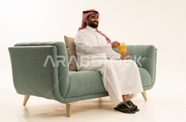 مشاهدة فيلم سينمائي، قضاء وقت مسلي و ممتع، الاستمتاع بتناول الفوشار، مفهوم التسليه والترفيه، بورتريه لرجل عربي سعودي خليجي يرتدي الشماغ والثوب التقليدي يجلس على الاريكة يحمل بيده علبة الفشار ويقوم بمشاهده الافلام، خلفيه بيضاء