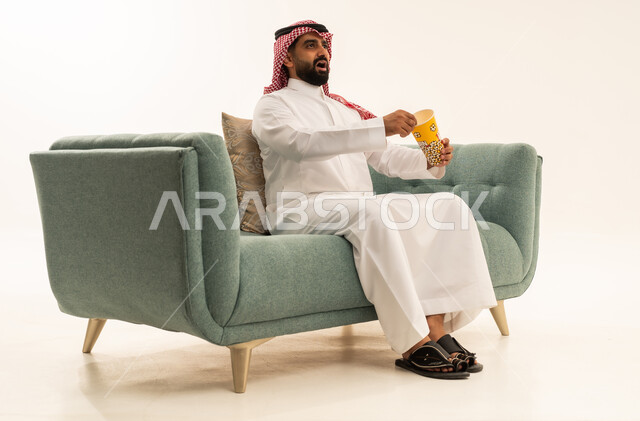 قضاء وقت مسلي و ممتع، الاستمتاع بتناول الفوشار، النظر بتعابير الاندهاش والاستغراب، مفهوم التسليه والترفيه، بورتريه لرجل عربي سعودي خليجي يرتدي الشماغ والثوب التقليدي يجلس على الاريكة يحمل بيده علبة الفشار ويقوم بمشاهده الافلام ، مشاهدة فيلم سينمائي ، خلفيه بيضاء