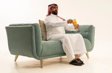 قضاء وقت مسلي و ممتع، الاستمتاع بتناول الفوشار، النظر بتعابير الاندهاش والاستغراب، مفهوم التسليه والترفيه، بورتريه لرجل عربي سعودي خليجي يرتدي الشماغ والثوب التقليدي يجلس على الاريكة يحمل بيده علبة الفشار ويقوم بمشاهده الافلام ، مشاهدة فيلم سينمائي ، خلفيه بيضاء