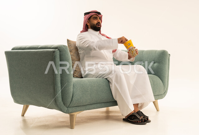 Enjoying popcorn, looking with expressions of amazement and wonder, the concept of amusement and entertainment, spending enjoyable and entertaining times, a portrait of an Arab Saudi Gulf man wearing a ghutrah and traditional thobe sitting on the couch holding a box of popcorn and watching movies, watching a cinematic film, white background.