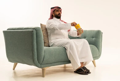 الاستمتاع بتناول الفوشار، النظر بتعابير الاندهاش والاستغراب، مفهوم التسليه والترفيه، قضاء أوقات ممتعة ومسلية، بورتريه لرجل عربي سعودي خليجي يرتدي الشماغ والثوب التقليدي يجلس على الاريكة يحمل بيده علبة الفشار ويقوم بمشاهده الافلام ، مشاهدة فيلم سينمائي ، خلفيه بيضاء