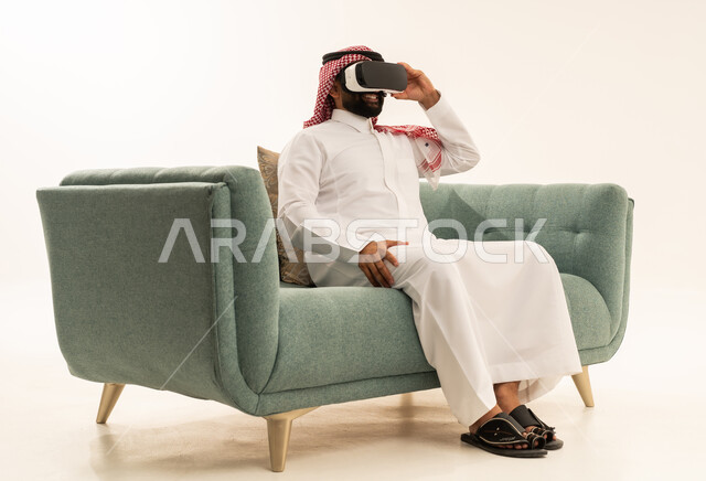 المتعة و التسلية بالمشاهدة عبر نظارة الواقع الافتراضي، التطور التقني والتكنولوجي، بورتريه لرجل عربي خليجي سعودي يرتدي الثوب التقليدي و الشماغ يجلس على الاريكة ينظر من خلال نظارات ثلاثية الأبعاد (الفي ار), الاندماج ومحاكاة العالم الخيالي والنظر الى الافلام الواقعيه، خلفيه بيضاء