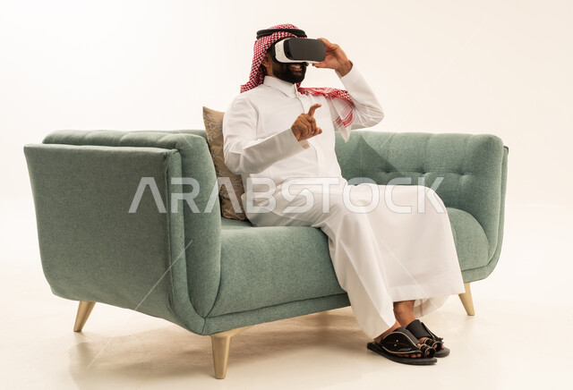 قضاء وقت الفراغ بشكل ممتع، الاشارة بإصبع السبابة إلى شيء ما، التفاعل والاندماج مع الواقع الافتراضي، استخدام الأجهزة التقنية الحديثة، رؤية المشاهد الخيالية، بورتريه لرجل عربي خليجي سعودي يرتدي الثوب التقليدي و الشماغ يجلس على الاريكة يضع نظارات الفي آر، خلفية بيضاء
