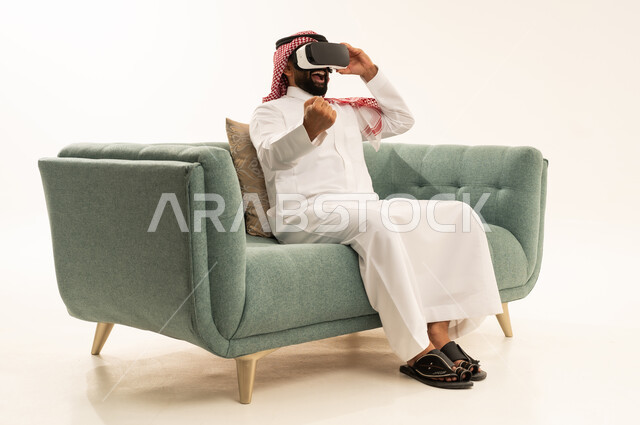 تعابير الفوز والانتصار، التطور التقني والتكنولوجي, المتعة و التسلية بالمشاهدة عبر نظارة الواقع الافتراضي، بورتريه لرجل عربي خليجي سعودي يرتدي الثوب التقليدي و الشماغ يجلس على الاريكة ينظر من خلال نظارات ثلاثية الأبعاد (الفي ار), الاندماج ومحاكاة العالم الخيالي والنظر الى الافلام الواقعيه، خلفيه بيضاء