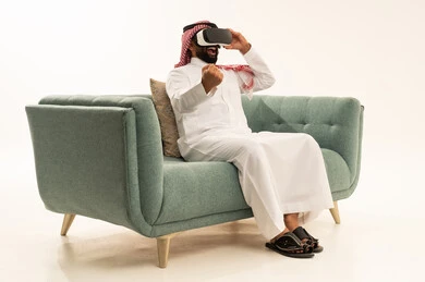 Expressions of victory and triumph, technological and technical development, enjoyment and entertainment through watching via virtual reality glasses, a portrait of a Saudi Gulf Arab man wearing traditional attire and a ghutrah sitting on a sofa looking through 3D glasses (VR), immersion and simulation of the imaginary world and watching realistic movies, white background.