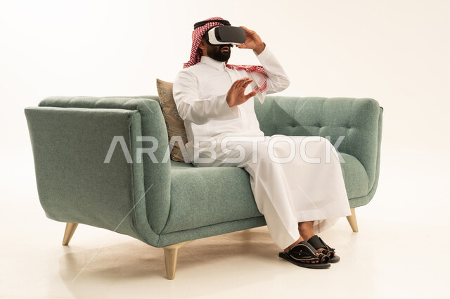Vision of imaginary scenes, raising the palm of the hand upwards with gestures of immersion and simulating the imaginary world, using modern technological devices, a portrait of a Saudi Arabian Gulf man wearing traditional attire and a ghutrah sitting on a sofa wearing VR glasses, enjoying leisure time, white background.