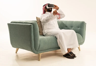 الاشارة بإصبع السبابة إلى شيء ما، التفاعل والاندماج مع الواقع الافتراضي، استخدام الأجهزة التقنية الحديثة، رؤية المشاهد الخيالية، بورتريه لرجل عربي خليجي سعودي يرتدي الثوب التقليدي و الشماغ يجلس على الاريكة يضع نظارات الفي آر، قضاء أوقات الفراغ بشكل ممتع، خلفية بيضاء