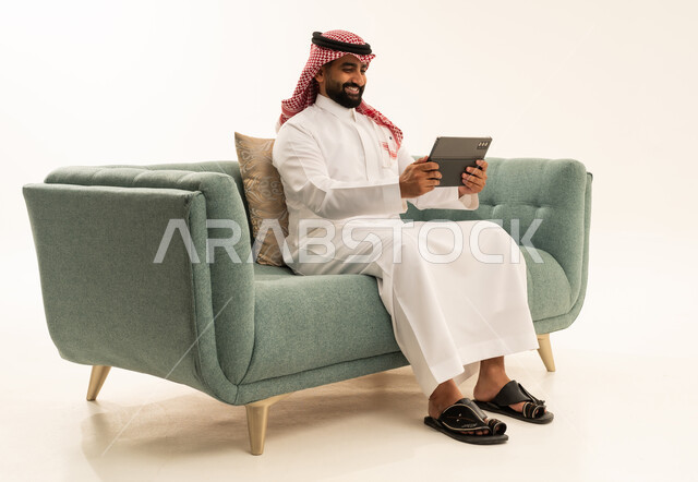 استخدام جهاز تقني حديث ومتطور، الدردشة مع الأصدقاء والأقارب عبر التابلت،ايماءات الفرح والسعادة،بورتريه لرجل عربي سعودي خليجي يرتدي الشماغ والثوب التقليدي يجلس على الاريكة يمسك بيده الجهاز اللوحي، خلفية بيضاء
