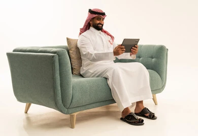 استخدام جهاز تقني حديث ومتطور، الدردشة مع الأصدقاء والأقارب عبر التابلت،ايماءات الفرح والسعادة،بورتريه لرجل عربي سعودي خليجي يرتدي الشماغ والثوب التقليدي يجلس على الاريكة يمسك بيده الجهاز اللوحي، خلفية بيضاء