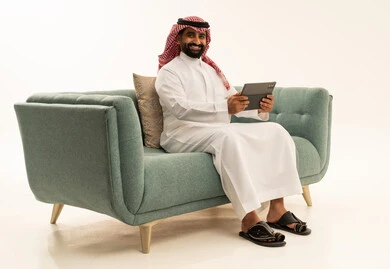 النظر للكاميرا بايماءات الفرح والسعادة، استخدام جهاز تقني حديث ومتطور، الدردشة مع الأصدقاء والأقارب عبر التابلت، بورتريه لرجل عربي سعودي خليجي يرتدي الشماغ والثوب التقليدي يجلس على الاريكة يمسك بيده الجهاز اللوحي، خلفية بيضاء