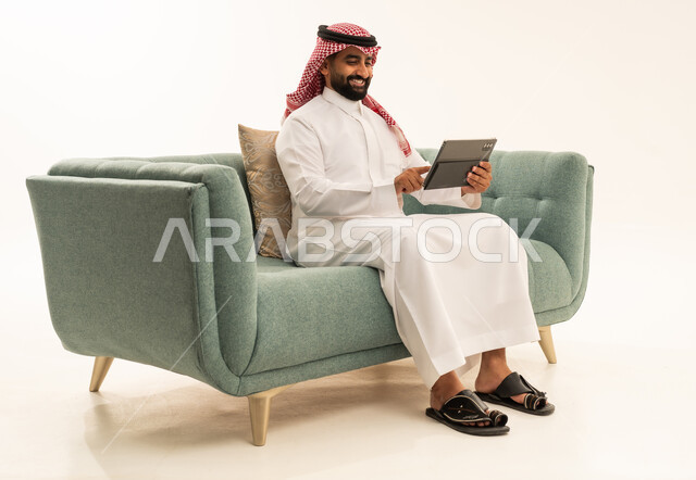 الدردشة مع الأصدقاء والأقارب عبر التابلت، استخدام جهاز تقني حديث ومتطور،ايماءات الفرح والسعادة،بورتريه لرجل عربي سعودي خليجي يرتدي الشماغ والثوب التقليدي يجلس على الاريكة يمسك بيده الجهاز اللوحي، خلفية بيضاء