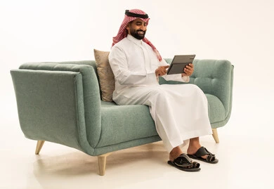 Chatting with friends and relatives via tablet, using a modern and advanced technological device, gestures of joy and happiness, a portrait of an Arab Saudi Gulf man wearing a ghutrah and traditional thobe sitting on the sofa holding the tablet in his hand, white background.