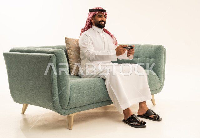 الاستمتاع بالتقنيات والتكنولوجيا الحديثة، إيماءات التفاعل والاندماج, بورتريه لرجل عربي سعودي خليجي يرتدي الشماغ والثوب التقليدي يجلس على الاريكة يمسك عصا التحكم ينظر إلى الكاميرا بتعابير السرور، مفهوم المتعة والتسلية، قضاء وقت ممتع في اللعب بألعاب الفيديو، خلفية بيضاء