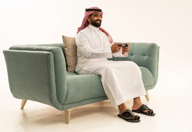 Enjoying modern technologies and technology, gestures of interaction and integration, a portrait of an Arab Saudi Gulf man wearing a ghutrah and traditional thobe sitting on a sofa holding a game controller looking at the camera with expressions of joy, the concept of fun and entertainment, having a good time playing video games, white background.