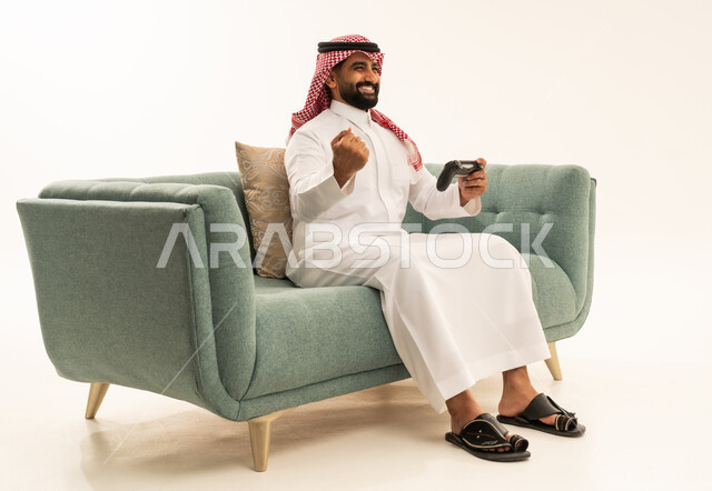 التفاعل والاندماج مع ألعاب البلايستيشن، تعابير الفوز والانتصار، تقنيات الكترونية حديثة ترفيهيه, بورتريه لرجل عربي سعودي خليجي يرتدي الشماغ والثوب التقليدي يجلس على الاريكة يحمل بيده عصا التحكم يقوم باللعب في ألعاب الفيديو، خلفية بيضاء