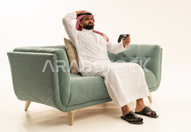 استخدام الألعاب الإلكترونية والأجهزة الحديثة، إيماءات الخسارة وخيبة الأمل، بورتريه لرجل عربي خليجي سعودي يرتدي الثوب الابيض و الشماغ يجلس على الاريكة يمسك عصا التحكم يضع يده على رأسه بتعابير الحسرة والحزن، التسلية والترفيه بالفيديو جيمز في وقت الفراغ، خلفية بيضاء