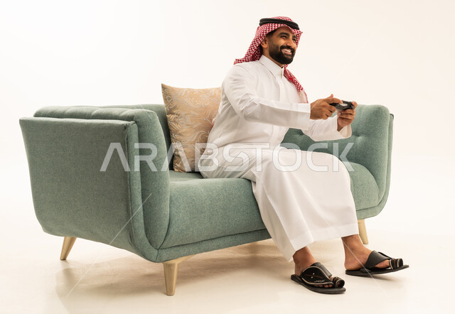 قضاء وقت ممتع في اللعب بألعاب الفيديو، إيماءات التفاعل والاندماج, بورتريه لرجل عربي سعودي خليجي يرتدي الشماغ والثوب التقليدي يجلس على الاريكة يمسك عصا التحكم ينظر إلى الكاميرا بتعابير السرور، مفهوم المتعة والتسلية، الاستمتاع بالتقنيات والتكنولوجيا الحديثة، خلفية بيضاء