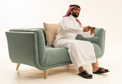 Spending enjoyable time playing video games, gestures of interaction and immersion, a portrait of an Arab Saudi Gulf man wearing a ghutrah and traditional thobe sitting on the sofa holding a game controller looking at the camera with expressions of joy, the concept of fun and entertainment, enjoying modern technologies and technology, white background.