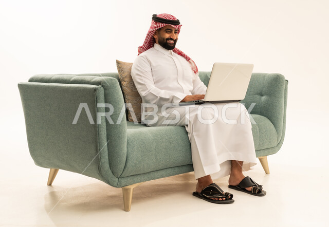 Remote work and completing required tasks, managing and organizing business affairs through technical devices, integrating work with technology, a portrait of an Arab Saudi Gulf man wearing a ghutrah and traditional thobe sitting on a sofa working on a laptop, using a modern and advanced technical device, white background.