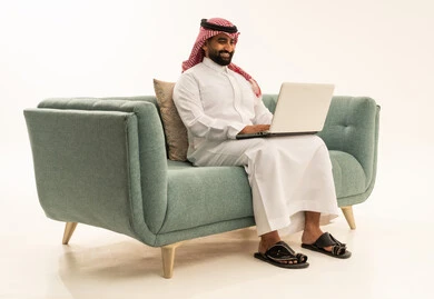 Remote work and completing required tasks, managing and organizing business affairs through technical devices, integrating work with technology, a portrait of an Arab Saudi Gulf man wearing a ghutrah and traditional thobe sitting on a sofa working on a laptop, using a modern and advanced technical device, white background.