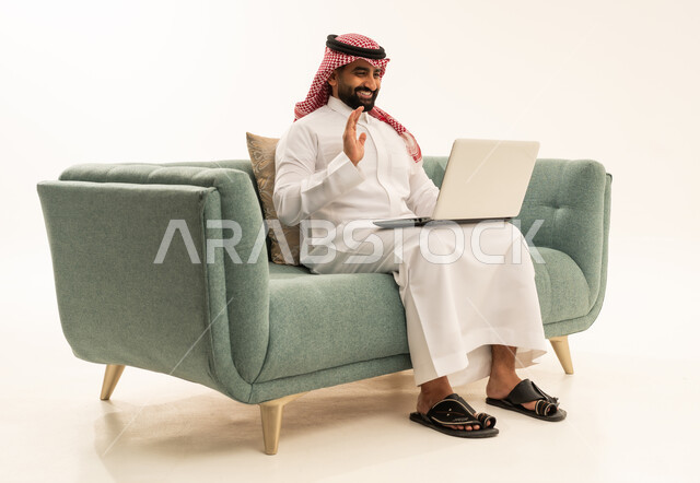 Raising the hand with expressions of peace and greeting, making a video call, working remotely, using advanced modern technology in professional life, a side portrait of a Saudi Arabian Gulf man wearing a ghutrah and traditional thobe sitting on a sofa working on a laptop, white background.