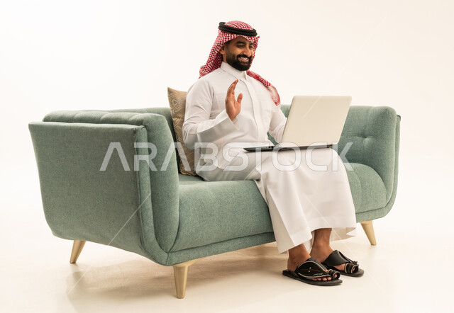 العمل عن بعد، رفع كف اليد بتعابير السلام و التحية، اجراء مكالمة فيديو، استخدام التقنية الحديثه المتطورة في الحياة العملية، بورتريه من الجانب لرجل عربي سعودي خليجي يرتدي الشماغ والثوب التقليدي يجلس على الاريكة يقوم بالعمل على الحاسب المحمول، خلفية بيضاء